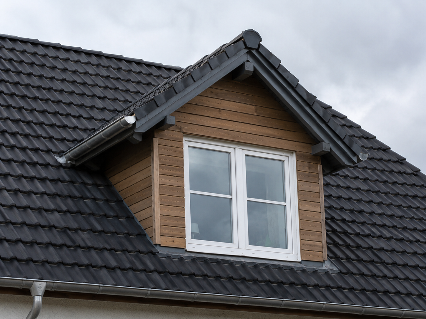 Satteldachgaube mit Holzverkleidung und weißem Fenster in einem dunkel eingedeckten Ziegeldach